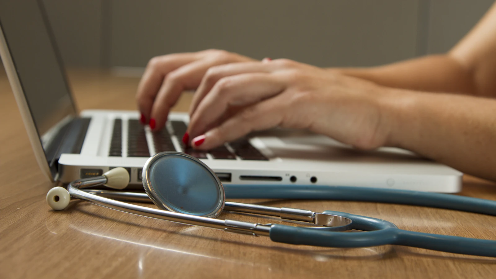 A doctor reviews blood results on a laptop, with a stethoscope on the desk.