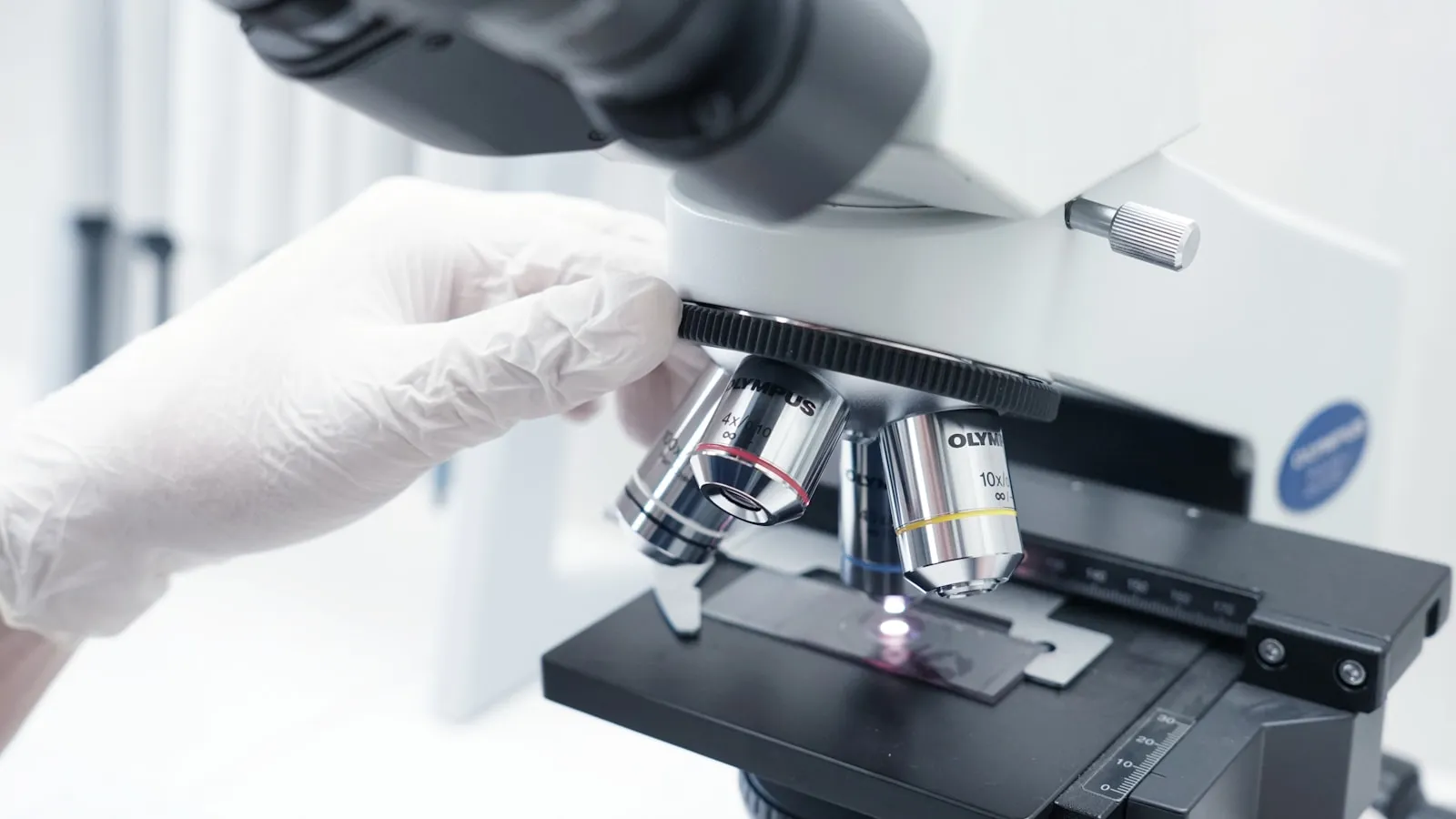 Laborant met witte handschoenen kijkt door een microscoop.