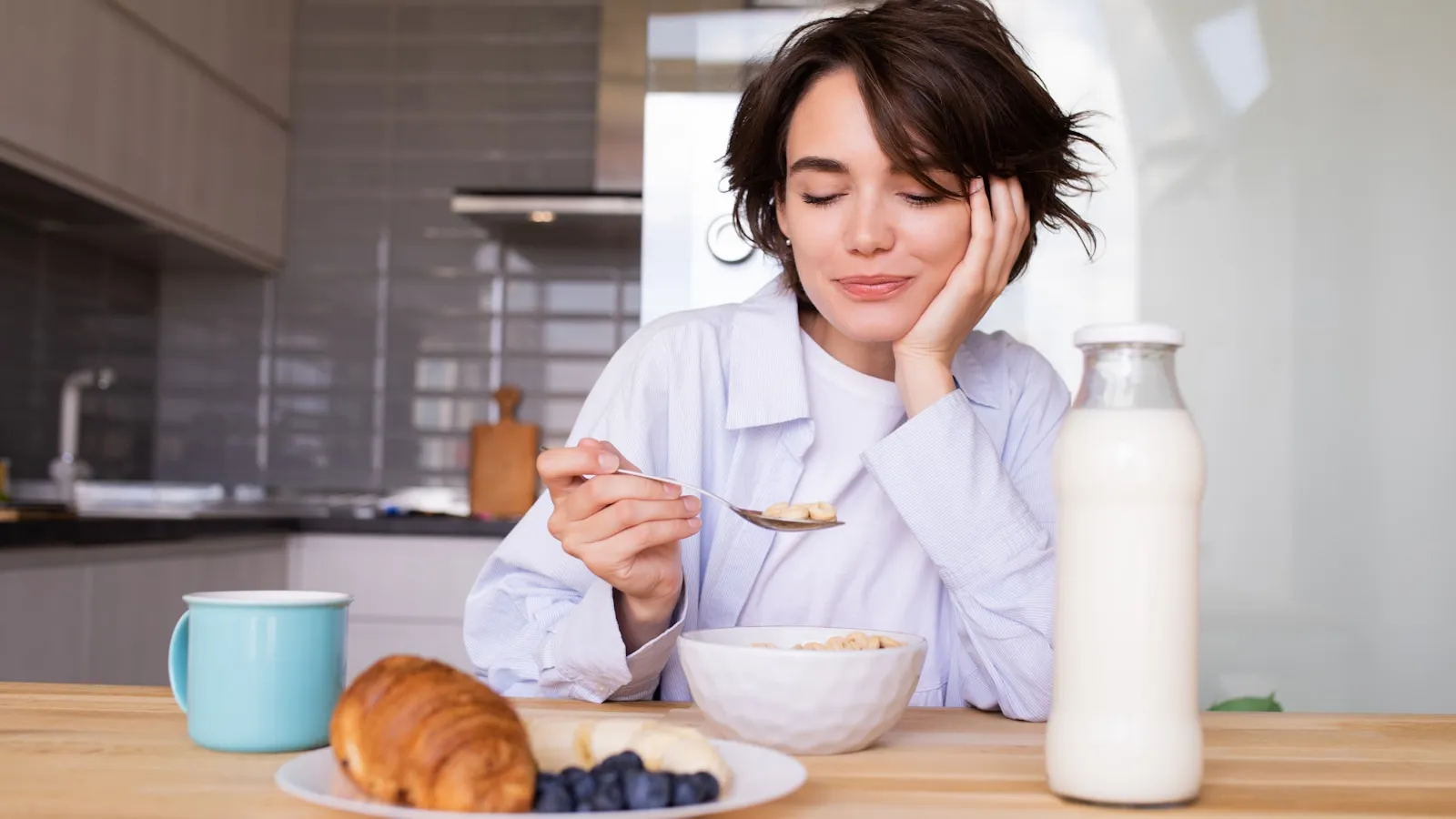 Vrouw aan de keukentafel met een kom ontbijt, een croissant en een fles melk.