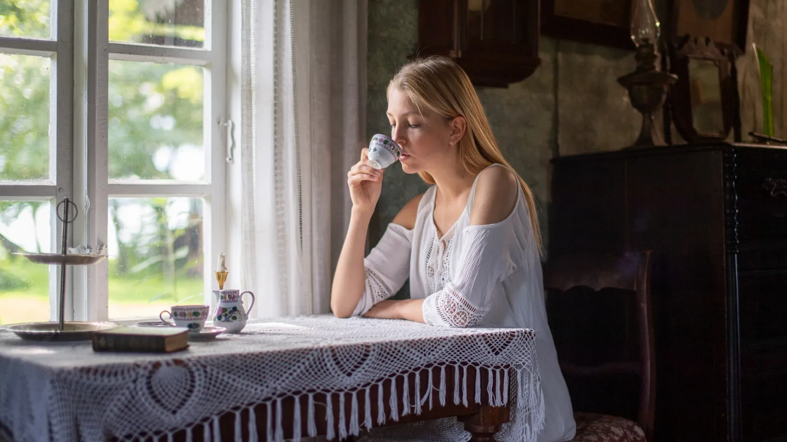 Een jonge vrouw drinkt thee bij het raam in daglicht.