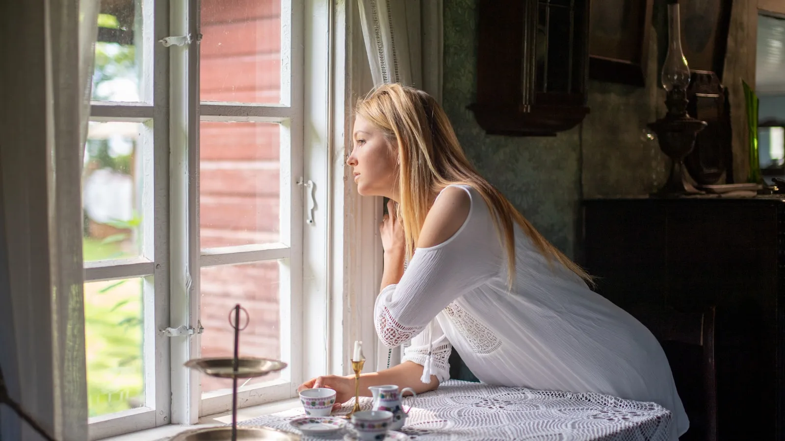 Een blonde vrouw in het wit kijkt uit een raam in het ochtendlicht, met een kopje thee op tafel.