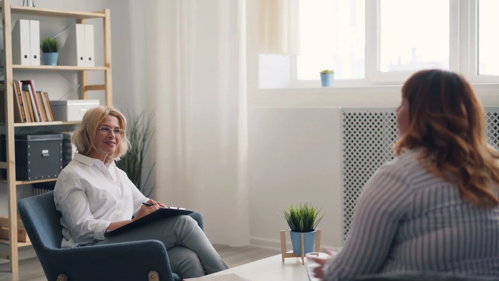 Oudere vrouw in gesprek met een zorgverlener in een lichte spreekkamer.